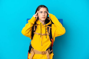 Mavi arka planda izole edilmiş büyük bir sırt çantası olan genç dağcı kadın şüpheleri ve düşünceleri var.