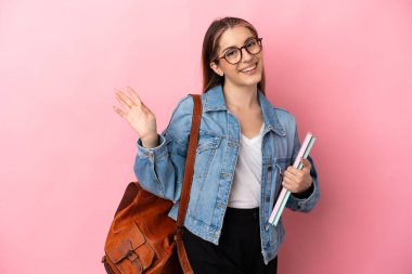 Beyaz tenli genç bayan pembe arka planda izole edilmiş, mutlu bir ifadeyle selamlanıyor.