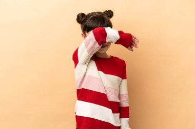 Bej arka planda izole edilmiş küçük beyaz kız gözlerini elleriyle kapatıyor.