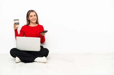 Dizüstü bilgisayarı olan, elinde kahve, elinde cep telefonu olan genç, beyaz bir kadın.