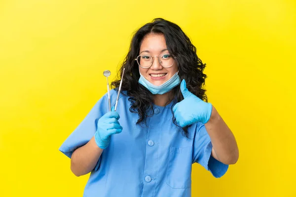 年轻的亚洲牙医拿着工具 带着大拇指站在孤立的背景下 因为发生了件好事 — 图库照片