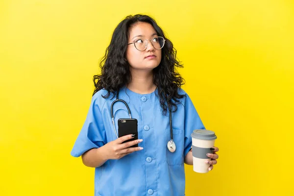 年轻的外科医生 亚洲女人 被隔离在黄色的背景下 一边拿着咖啡 一边想着什么 一边拿着手机 — 图库照片