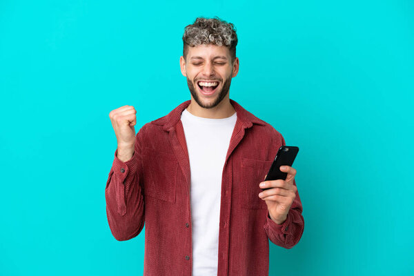 Young handsome caucasian man isolated on blue background using mobile phone and doing victory gesture