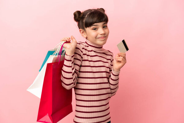 Маленькая белая девочка, изолированная на розовом фоне, держащая сумки с покупками и кредитку