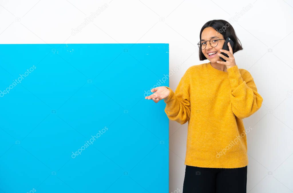 Young mixed race woman with a big blue placard isolated on white background keeping a conversation with the mobile phone with someone