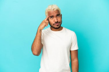 Genç Kolombiyalı yakışıklı adam mavi arka planda izole edilmiş. Delilik jesti yapıyor. Parmağını kafaya koyuyor.