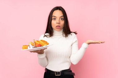 Soyutlanmış pembe arka planda waffle tutan esmer bir kadın şüpheli hareketlerde bulunuyor.
