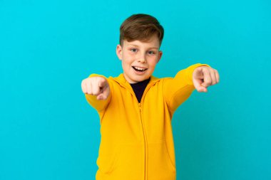 Küçük kızıl saçlı çocuk mavi arka planda izole edilmiş şaşırmış ve ön tarafı işaret ediyor.