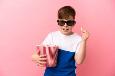 Küçük kızıl saçlı çocuk pembe arka planda 3 boyutlu gözlüklü ve elinde patlamış mısır dolu bir kova tutuyor.