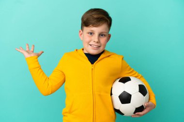 Kırmızı saçlı çocuk mavi arka planda izole bir şekilde futbol oynuyor.