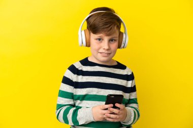 Küçük kızıl saçlı çocuk sarı arka planda izole edilmiş. Cep telefonuyla müzik dinliyor ve ön tarafa bakıyor.