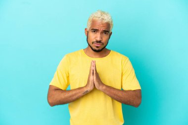 Mavi arka planda izole edilmiş genç Kolombiyalı yakışıklı adam avuç içlerini bir arada tutuyor. İnsan bir şey ister.