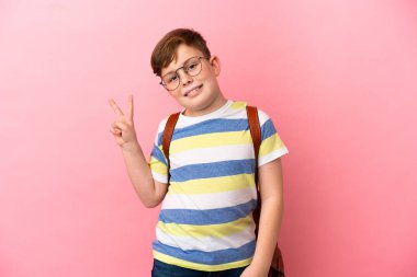 Küçük kızıl saçlı beyaz çocuk pembe arka planda izole edilmiş gülümsüyor ve zafer işareti gösteriyor.