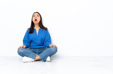 Genç melez bir kadın, beyaz arka planda izole bir şekilde yerde oturuyor ve şaşırmış bir şekilde yukarı bakıyor.