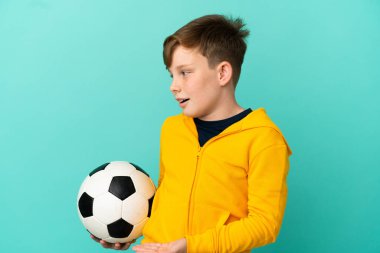 Mavi arka planda futbol oynayan kızıl saçlı çocuk şaşırmış bir ifadeyle yan tarafa bakıyor.