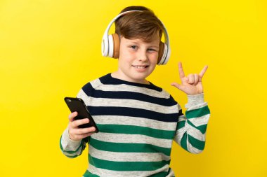 Küçük kızıl saçlı çocuk sarı arka planda izole edilmiş gezici bir rock hareketi yaparak müzik dinliyor.