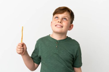 Beyaz arka planda izole edilmiş diş fırçası tutan küçük kızıl çocuk gülümserken yukarı bakıyor.