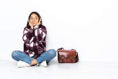 Genç bir bayan öğrenci, hayal kırıklığına uğramış ve kulaklarını kapatmış bir şekilde yerde oturuyor.