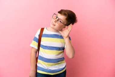 Küçük kızıl saçlı beyaz çocuk pembe arka planda izole edilmiş bir şekilde elini kulağına koyarak bir şeyler dinliyor.