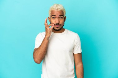 Mavi arka planda izole edilmiş genç Kolombiyalı yakışıklı bir adam sessizlik işareti gösteriyor.