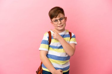 Pembe arka planda izole edilmiş küçük kızıl saçlı çocuk bir ürünü sunmak için kenarı işaret ediyor.