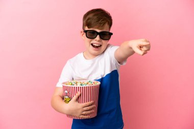 Küçük kızıl saçlı çocuk pembe arka planda 3 boyutlu gözlüklerle izole edilmiş ve elinde patlamış mısır kovası tutuyordu.