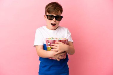 Pembe arka planda izole edilmiş küçük kızıl saçlı çocuk 3D gözlüklü ve elinde koca bir kova patlamış mısırla şaşırdı.