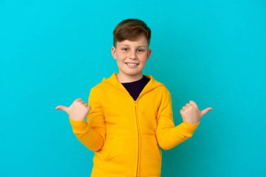 Küçük kızıl saçlı çocuk mavi arka planda izole edilmiş. Baş parmakları havada ve gülümsüyor.