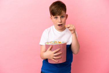 Pembe arka planda izole edilmiş küçük kızıl çocuk elinde koca bir kova patlamış mısır tutuyor.