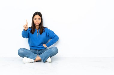 Genç melez kadın, beyaz arka planda izole bir şekilde yerde oturuyor. Ciddi bir ifadeye sahip birini sayıyor.