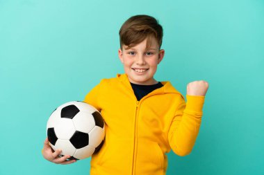 Mavi arka planda futbol oynayan kızıl saçlı çocuk galibiyeti kutluyor.