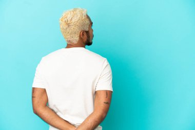 Genç Kolombiyalı yakışıklı adam mavi arka planda izole edilmiş ve arkasına bakıyor.