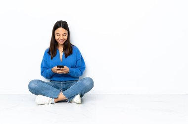 Genç melez kadın, beyaz arka planda izole bir şekilde yerde oturuyor ve cep telefonuyla bir mesaj gönderiyor.