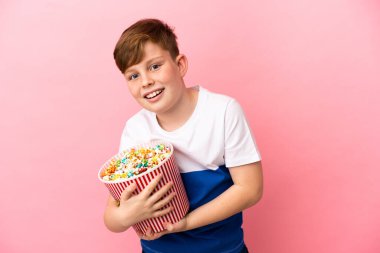 Pembe arka planda izole edilmiş küçük kızıl çocuk elinde koca bir kova patlamış mısır tutuyor.