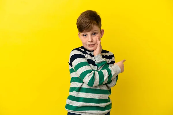 一个红头发的小男孩在黄色的背景上被隔离起来 指着侧面展示了一种产品 并低声说了些什么 — 图库照片