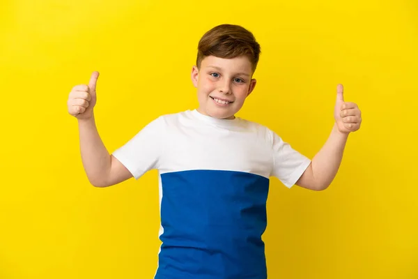 小さな赤い頭の男の子は黄色の背景に隔離されている親指を上げるジェスチャー — ストック写真