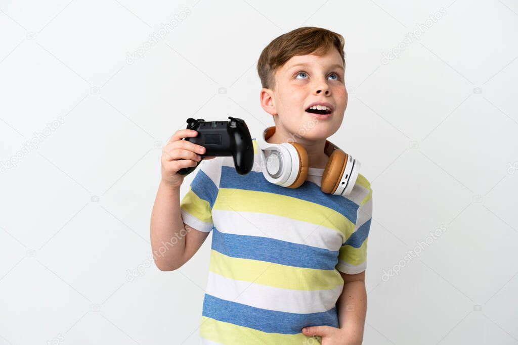 Little redhead boy holding a game pad isolated on white background looking up and with surprised expression