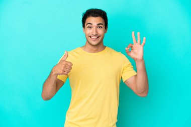 İzole edilmiş mavi arka planda beyaz, yakışıklı bir adam tamam işareti ve baş parmak hareketi gösteriyor.