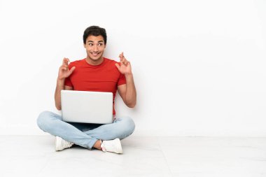 Caucasian handsome man with a laptop sitting on the floor with fingers crossing