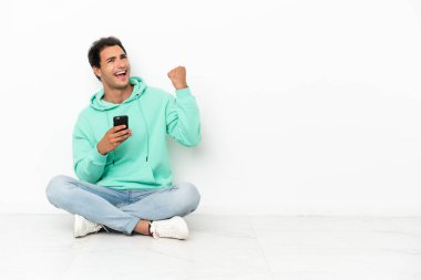 Caucasian handsome man sitting on the floor with phone in victory position