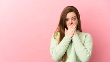 İzole edilmiş pembe arka planı olan genç bir kız elleriyle ağzını kapatıyor.