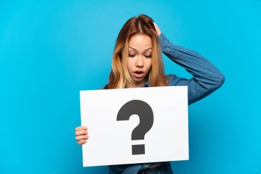İzole edilmiş mavi arkaplanı olan genç bir kız soru işareti sembollü bir levha tutuyor ve şaşırmış bir ifade var.