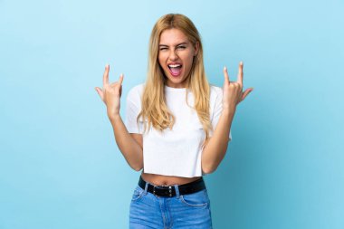 Genç Uruguaylı sarışın kadın izole edilmiş mavi arka planda boynuz hareketi yapıyor.