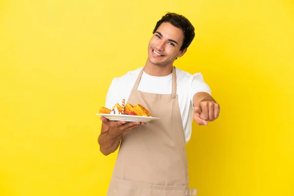 独立した黄色の背景の上にワッフルを保持するレストランウエイター幸せな表情で前を指す — ストック写真