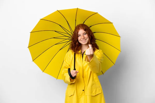 ティーンRedhead女の子雨防止コートと傘隔離上のホワイトバックグランド作るお金ジェスチャー — ストック写真