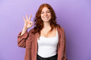 İzole edilmiş mor arka planda genç bir kızıl kız parmaklarıyla tamam işareti gösteriyor.