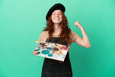 Yeşil arka planda izole edilmiş palet tutan kızıl saçlı genç sanatçı güçlü bir jest yapıyor.