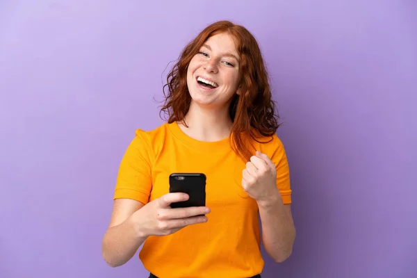 Подростковая Рыжая Девушка Изолированном Фиолетовом Фоне Телефоном Победной Позиции — стоковое фото