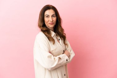 Orta yaşlı beyaz kadın pembe arka planda izole edilmiş, kolları bağlı ve ileriye bakıyor.