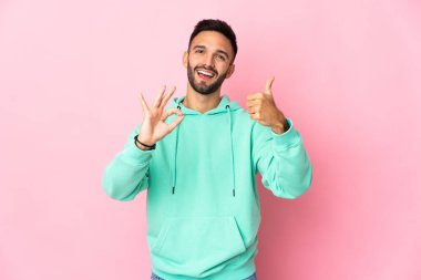 Pembe arka planda izole edilmiş genç beyaz bir adam tamam işareti ve baş parmak hareketi gösteriyor.